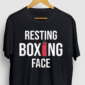 Könnte beinhalten: Schwarzes T-Shirt mit weißem Text "Resting Boxing Face" und einer roten Boxsackgrafik.
