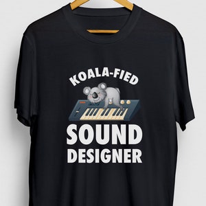 Op de afbeelding: Zwart T-shirt met witte tekst die "Koala-fied Sound Designer" luidt. Een cartoonkoala slaapt op een synthesizertoetsenbord.
