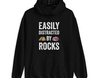 Puede incluir: Sudadera con capucha negra con un gráfico blanco que dice "Easily Distracted by Rocks" con dos pequeñas ilustraciones de rocas.