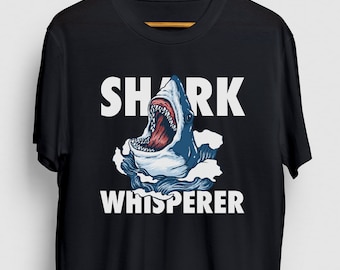 May include: Black t-shirt with a graphic of a blue and white shark with an open mouth, teeth visible. The text "SHARK WHISPERER" is printed above and below the shark illustration in white.