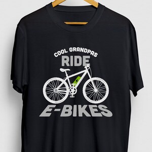 Puede incluir: Camiseta negra con texto blanco que dice "Cool Grandpas Ride E-Bikes". Una silueta blanca de una bicicleta eléctrica con una batería verde está en el centro de la camiseta.