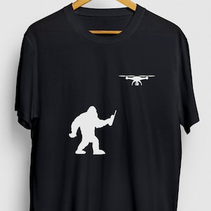 May include: A black t-shirt with a white silhouette of a Bigfoot holding a remote control and a drone flying above him.