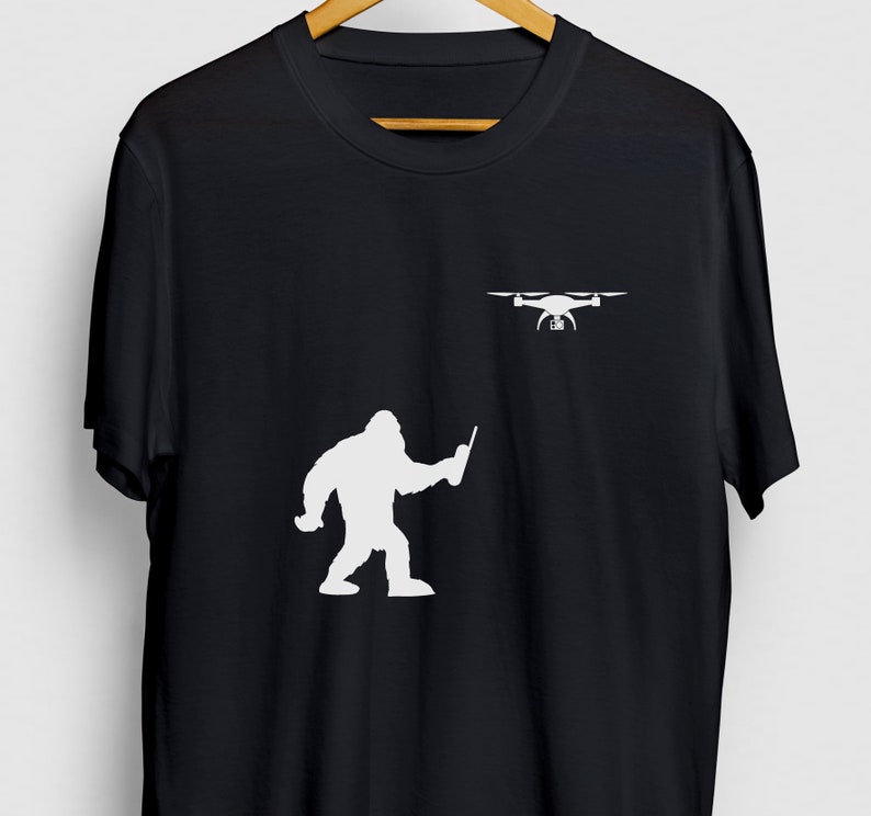 May include: A black t-shirt with a white silhouette of a Bigfoot holding a remote control and a drone flying above him.
