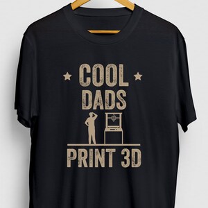 May include: Black t-shirt with a brown graphic that says "Cool Dads Print 3D". The graphic includes a star on each side of the word "Cool" and a silhouette of a person standing next to a 3D printer.