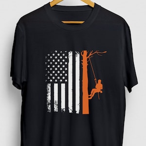 以下が含まれることがあります： アメリカ国旗のデザインが施された黒のTシャツ。木に登る人のシルエットが描かれている。