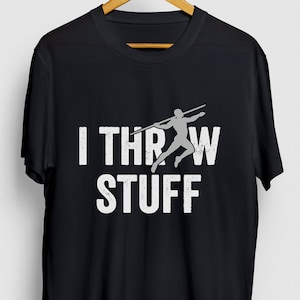 Puede incluir: Camiseta negra con el texto blanco "I THROW STUFF" y un gráfico gris de una persona lanzando una jabalina. La camiseta está colgada en una percha de madera.