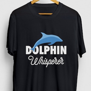 Pode incluir: Camiseta preta com um gráfico de golfinho azul e o texto "Dolphin Whisperer" em branco.