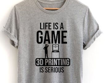 Könnte beinhalten: Ein graues T-Shirt mit schwarzem Text, der "Life is a game 3D printing is serious" lautet. Der Text ist mittig auf dem Shirt platziert und von einer kleinen Illustration einer Person umgeben, die eine 3D-Druckmaschine betrachtet.