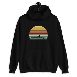Puede incluir: Sudadera con capucha negra con un gráfico de puesta de sol retro que presenta la silueta de una persona remando un bote.