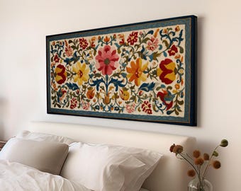 May include: A framed floral tapestry with a cream background and a dark blue border. The tapestry features a vibrant array of flowers in shades of pink, yellow, orange, and red, with green and blue foliage. The artwork is mounted above a bed.