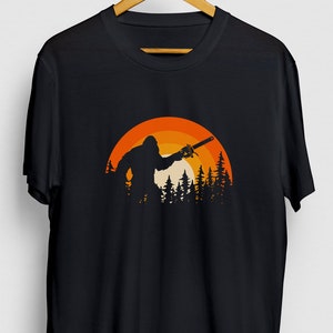 Op de afbeelding: Zwart T-shirt met een retro-stijl grafische afbeelding van Bigfoot die een kettingzaag vasthoudt voor een zonsondergang en bomen.