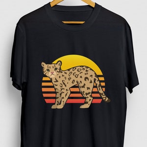 Może przedstawiać: Czarny t-shirt z grafiką retro zachodu słońca przedstawiającą brązowego i czarnego ocelota stojącego przed żółtym słońcem z pomarańczowymi i czerwonymi paskami.
