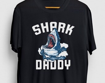 May include: Black t-shirt with a white graphic of a shark with its mouth open and the text "SHARK DADDY" underneath.