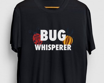 Può includere: Maglietta nera con testo bianco che recita "BUG WHISPERER". Il testo è circondato da due insetti dei cartoni animati, uno rosso e uno giallo.
