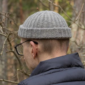 Könnte beinhalten: Graue Rippstrick-Beanie. Der Hut besteht aus einem weichen, strukturierten Material und hat eine enge Passform. Die Beanie ist eine neutrale Farbe und kann in verschiedenen Umgebungen getragen werden. Der Hintergrund ist eine verschwommene Waldszene.