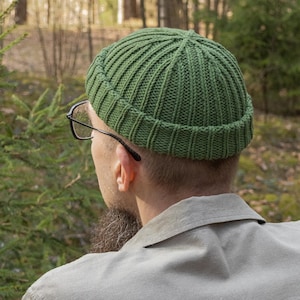 May include: A green ribbed knit beanie. The hat is a solid color and has a folded brim. The person is wearing glasses and a light-colored shirt. The background is a blurred forest scene.