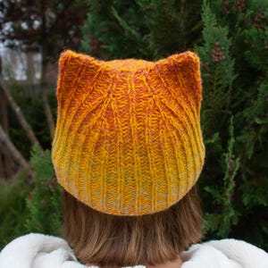May include: A knitted cat ear hat in shades of yellow, orange, and red. The hat is ribbed and has a rounded top with pointed ears. The person wearing the hat is wearing a white fluffy jacket.
