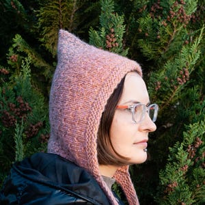 May include: A knitted pink hood with a pointed top. The hood is made of a textured yarn and is worn over the head. The person in the image is wearing glasses with orange frames and a black jacket. The background is a green bush.