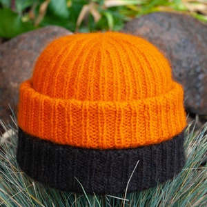 May include: A two-toned knit beanie featuring a vibrant orange upper section and a dark charcoal black lower brim. The hat is constructed with a vertical ribbed texture throughout, showcasing a soft, fuzzy yarn finish and a folded cuff design.