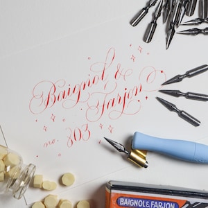 May include: A calligraphy arrangement with a white paper displaying red script: "Baigniol & Farjon no. 303". Calligraphy nibs, a blue pen holder, a jar of wax seals, and a box with the brand name are also present.