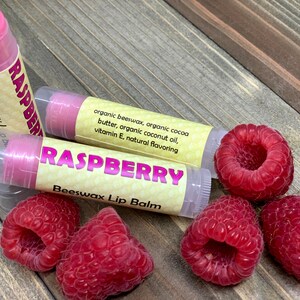 May include: Three pink raspberry flavored beeswax lip balms with yellow labels and the words "Raspberry Beeswax Lip Balm" on a wooden surface with four fresh raspberries.