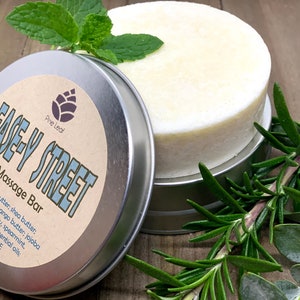 May include: A silver tin with a label that reads "Ease-Y Street Massage Bar" and lists ingredients including cocoa butter, shea butter, coconut oil, mango butter, jojoba oil, rosemary, spearmint, eucalyptus essential oils, and vitamin E. The tin contains a white, round massage bar.