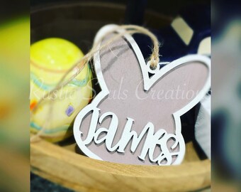 May include: A light brown wooden bunny-shaped ornament with the name "James" written in white lettering. The ornament is hanging from a string.