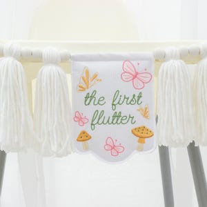 May include: White high chair banner with large white tassels and a decorative banner. The banner has the text "the first flutter" with embroidered butterflies and mushrooms. The banner is attached to a high chair.