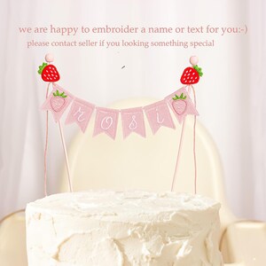 Embroidered name cake topper on pink cotton flags with strawberries on each end. Personalized topper for berry first birthday celebration.