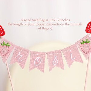 Embroidered name cake topper on pink cotton flags with strawberries on each end. Personalized topper for berry first birthday celebration.