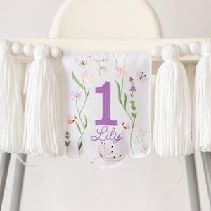 Pu&ograve; includere: Un banner bianco per seggiolone con nappe bianche e perline di legno. Il banner presenta un design floreale con il numero 1 viola, il nome "Lily" e illustrazioni di fiori e farfalle.