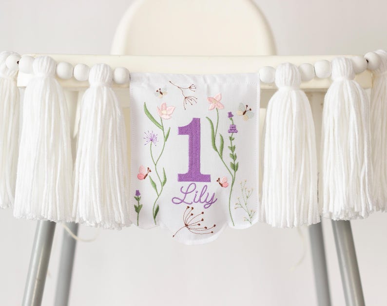 Pu&ograve; includere: Un banner bianco per seggiolone con nappe bianche e perline di legno. Il banner presenta un design floreale con il numero 1 viola, il nome "Lily" e illustrazioni di fiori e farfalle.