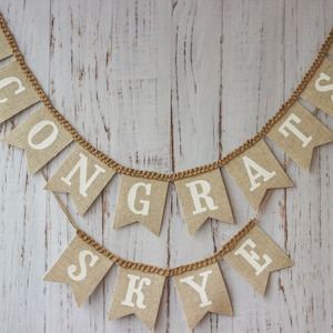 May include: A burlap banner with the words "CONGRATS SKYE" in white letters. The banner is decorated with graduation caps at each end.