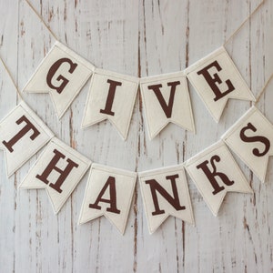 May include: A white fabric banner with brown letters spelling "GIVE THANKS" hanging on a white wood background.