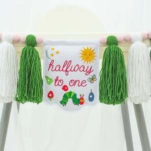 May include: A white banner with the text "halfway to one" and illustrations of a sun, caterpillar, and fruit. The banner is adorned with green and white tassels and pink wooden beads, designed for a baby's high chair.