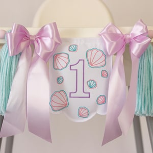 White embroidered mermaid high chair banner with seashells, number one, and lavender bows for a girl’s first birthday.