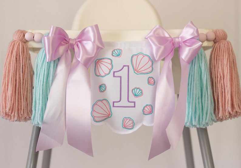 White embroidered mermaid high chair banner with seashells, number one, and lavender bows for a girl’s first birthday.
