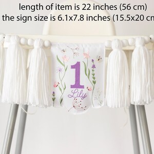 Pu&ograve; includere: Striscione bianco per seggiolone con nappe e ricami floreali. Lo striscione &egrave; lungo 56 cm e presenta il numero "1" in viola, il nome "Lily" e un motivo floreale. La dimensione del cartello &egrave; 15,5x20 cm.