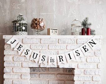 May include: A white brick fireplace mantel decorated with a banner that reads "JESUS is RISEN". The mantel also features decorative birdcages, a glass jar with nuts, a wooden calendar, and red candles.