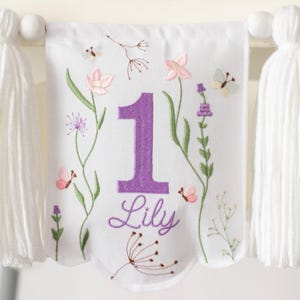 Pu&ograve; includere: Striscione di tessuto bianco ricamato con un grande numero 1 viola e il nome "Lily". Lo striscione presenta ricami floreali e di farfalle in rosa, verde e viola. Nappe bianche pendono su entrambi i lati.