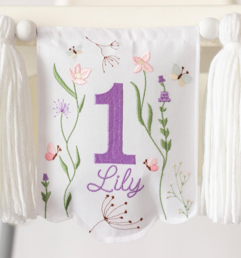 Pu&ograve; includere: Striscione di tessuto bianco ricamato con un grande numero 1 viola e il nome "Lily". Lo striscione presenta ricami floreali e di farfalle in rosa, verde e viola. Nappe bianche pendono su entrambi i lati.