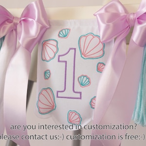 White embroidered mermaid high chair banner with seashells, number one, and lavender bows for a girl’s first birthday.