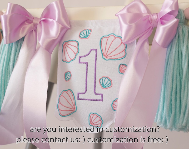 White embroidered mermaid high chair banner with seashells, number one, and lavender bows for a girl’s first birthday.