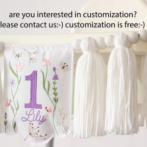 Pu&ograve; includere: Striscione in tessuto bianco con ricami floreali e farfalle, un grande numero 1 viola e il nome "Lily". Lo striscione &egrave; adornato con nappe bianche e perline di legno. Il testo in alto recita "are you interested in customization? please contact us:-) customization is free:-)"