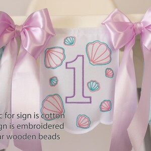 White embroidered mermaid high chair banner with seashells, number one, and lavender bows for a girl’s first birthday.