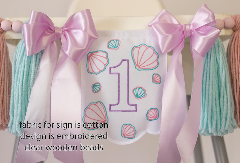 White embroidered mermaid high chair banner with seashells, number one, and lavender bows for a girl’s first birthday.