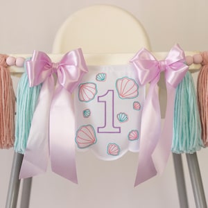 White embroidered mermaid high chair banner with seashells, number one, and lavender bows for a girl’s first birthday.