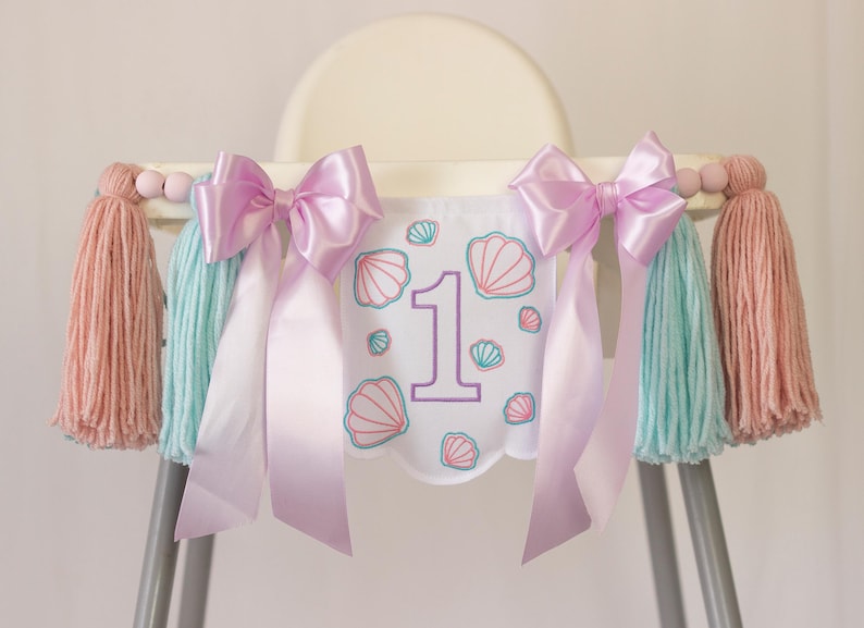 White embroidered mermaid high chair banner with seashells, number one, and lavender bows for a girl’s first birthday.