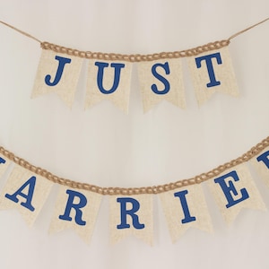 May include: A two-tiered banner made of light beige, textured fabric pennants strung on brown twine. The top row displays the word JUST in bold blue capital letters, and the bottom row displays the word MARRIED in bold blue capital letters.