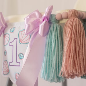 White embroidered mermaid high chair banner with seashells, number one, and lavender bows for a girl’s first birthday.